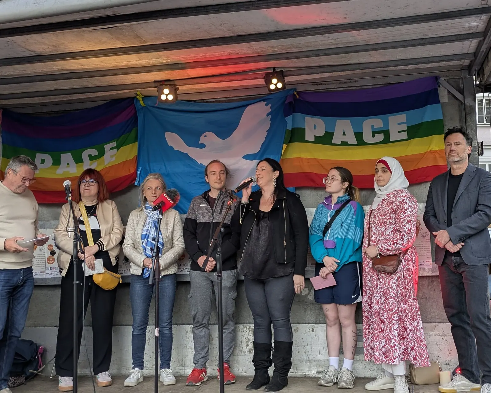 Neun Menschen stehen nebeneinander auf der Bühne vor Fahnen mit Friedenstaube und dem Wort Pace