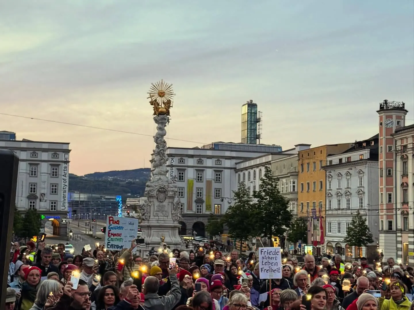 Abenddämmerung über dem Hauptplatz mit der Dreifaltigkeitssäule, davor eine große Menge mit Lichtern und Schildern